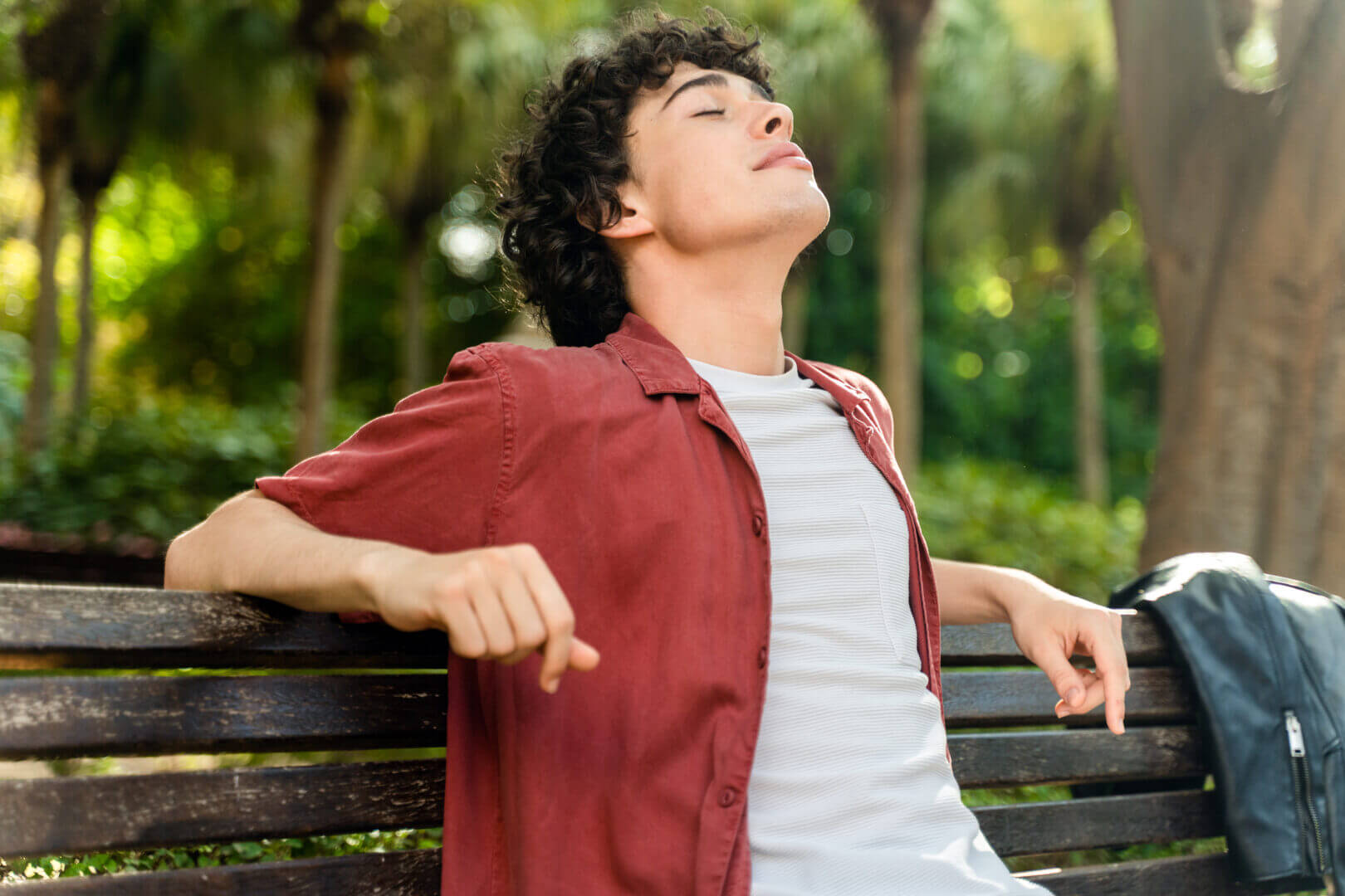 Happy curly man relaxing with closed eyes while sitting on the bench at the park