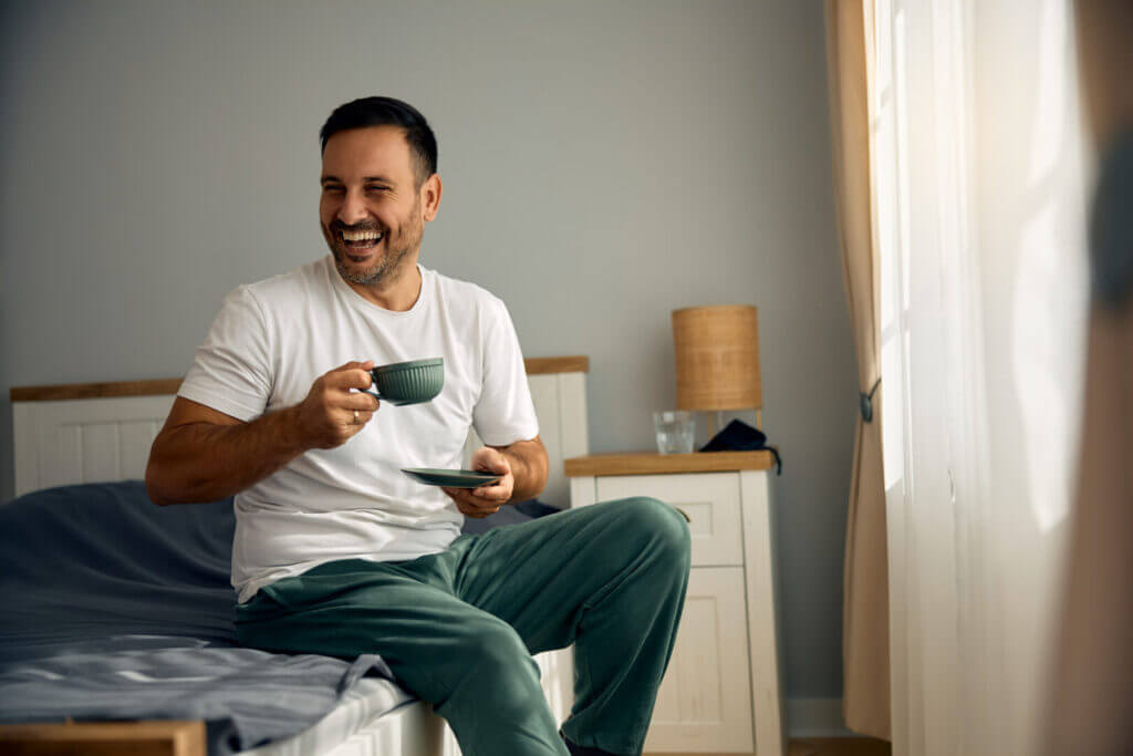 Happy man having cup of coffee in bedroom in the morning.